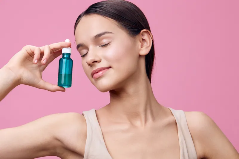 Young woman holding a cosmetic serum on a pink background, showcasing healthy skincare and beauty, with a serene expression and natural elegance