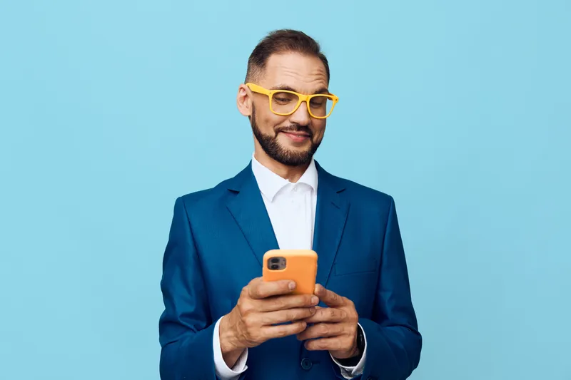 A professional man in a blue suit stands against a vivid blue backdrop, focused on his phone, conveying confidence, curiosity, and calm engagement.