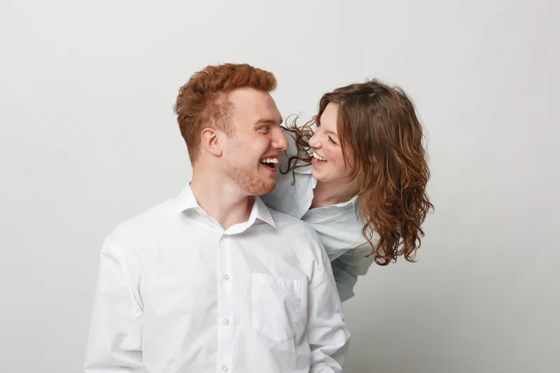 Happy young couple with curly hair laughing and looking at each other on white background. Concept of love, friendship, joy, and positive emotions in casual lifestyle.