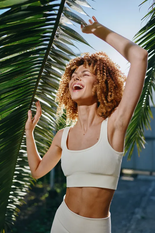young woman with curly hair enjoys sunshine outdoors, wearing white sportswear near palm leaves, radiant and joyful expression in natural light, summer atmosphere