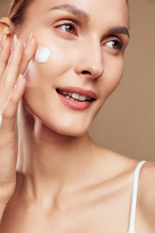 Young woman applies moisturizing cream on clean face with gentle touch smiling with natural skin care routine isolated on beige background with positive fresh expression.