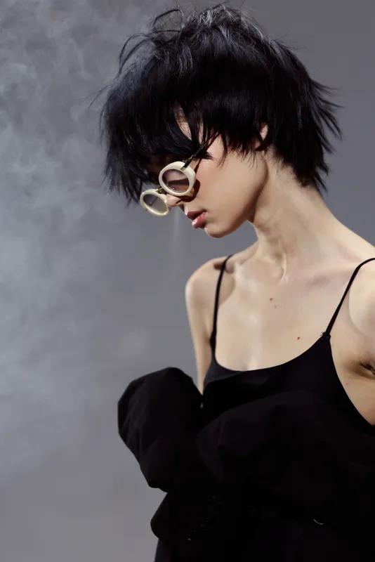 Stylish woman with short black hair and glasses wearing a black outfit posing gracefully with soft lighting and muted smoke background.