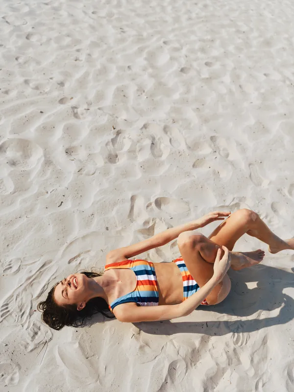 Sunlit beach scene with a reclining woman in a striped bikini resting on soft white sand, capturing a peaceful moment of sun, warmth, and carefree summer relaxation.