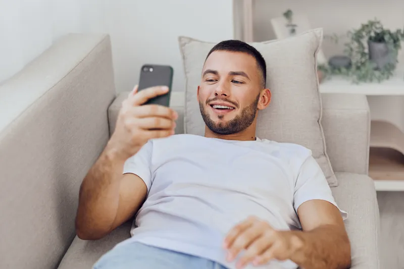 Engaging man relaxing on a couch, looking at his smartphone with a smile, showcasing modern lifestyle and technology. Perfect for contemporary digital themes.