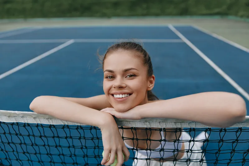 Smiling female tennis player leaning on the net, showcasing joy and confidence on a blue tennis court background Perfect for sports and fitness themes