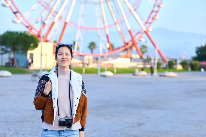 Camera in hand, woman smiles at a bustling amusement park, outdoor scenery with a rollercoaster in the background, casual jacket, photography and travel vibe on a sunny day.