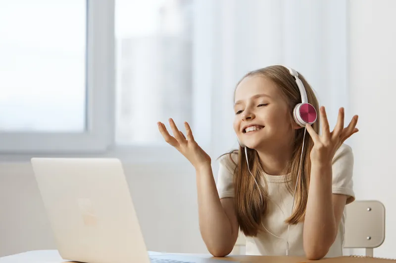 Happy girl with headphones enjoying music while sitting at a laptop in a bright, minimalistic room, expressing joy and engagement in a modern, youthful atmosphere