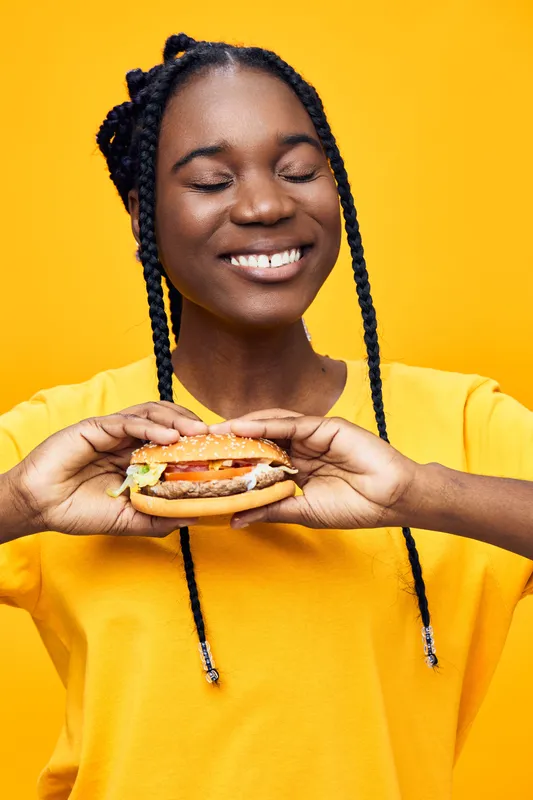 Happy young Black woman joyfully holding a delicious hamburger in front of a vibrant yellow background, wearing a bright yellow shirt, celebrating healthy eating
