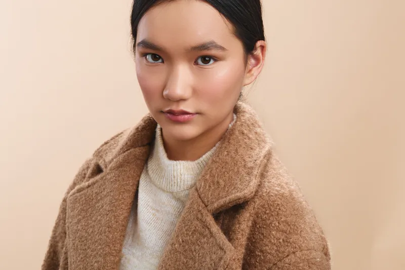 Asian woman with a calm expression poses in a soft beige studio backdrop, wearing a tan wool coat and white knit sweater, presenting a poised, stylish portrait.