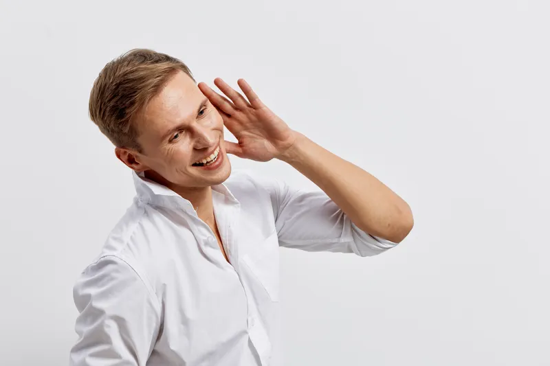 smiling man wearing white shirt listening gesture hold hand near ear isolated on light background mental health and gadgets concept