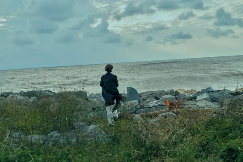 Figure in dark jacket on a rocky shore gazing at the sea, tranquil horizon and cloudy sky, coastal landscape, wild vegetation, and distant water line, conveying solitude, reflection, and outdoor