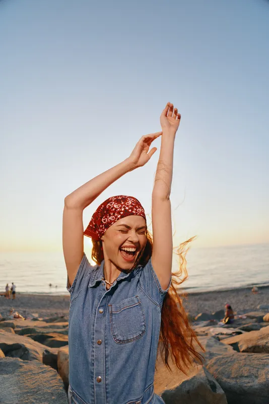joyful woman dancing at beach wearing denim dress and red bandana with sunset over calm ocean in background