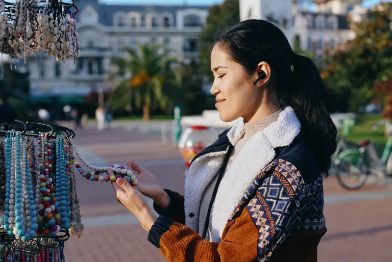 Female shopper examines colorful bead jewelry at an outdoor market stall, wearing a patterned jacket and scarf, sunlit urban plaza with vibrant accessories