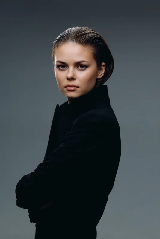 Serious woman in black turtleneck looking at camera with folded arms, neutral background, moody portrait style.