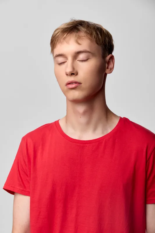 Calm youth, closed eyes, in a red shirt, conveys relaxation and stillness for wellness, mindfulness, and tranquil lifestyle themes in a studio setting. Natural tones and soft lighting enhance mood,