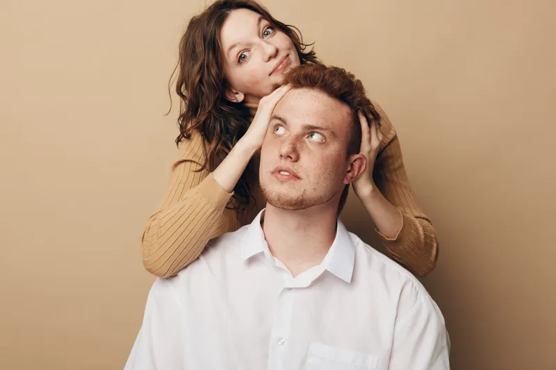 Young woman with brown hair gently touches head of redheaded man in white shirt. Both look thoughtful against beige background. Concept of care, connection, and emotional support.