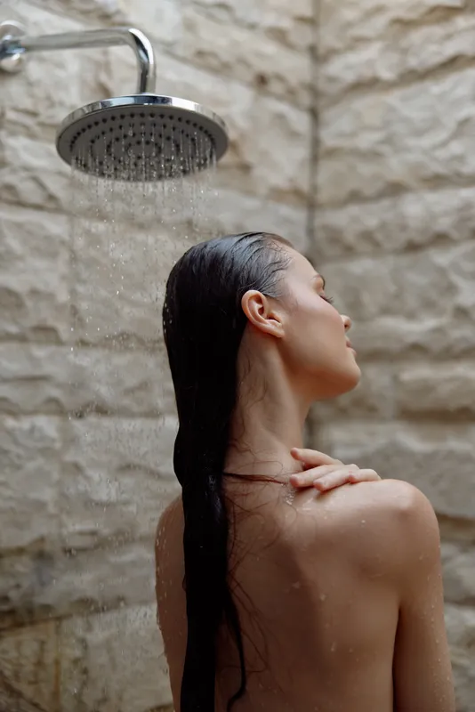 woman showering outdoors with wet hair under rain showerhead on textured stone wall background, natural light and relaxed mood