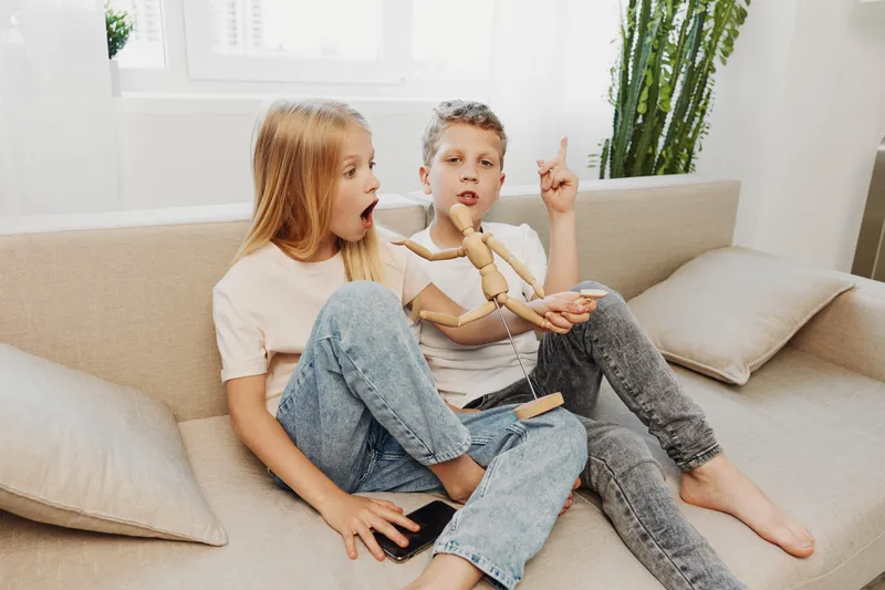 Two children sitting on sofa indoors playing with wooden mannequin, expressing surprise and curiosity in bright modern living room setting.