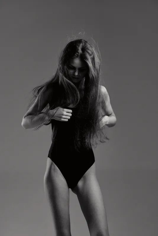 Black and white portrait of a young woman with long hair wearing a black bodysuit in a minimalistic studio setting.