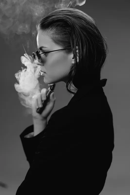Black and white portrait of a woman vaping with glasses in a dark outfit exhaling smoke indoors.