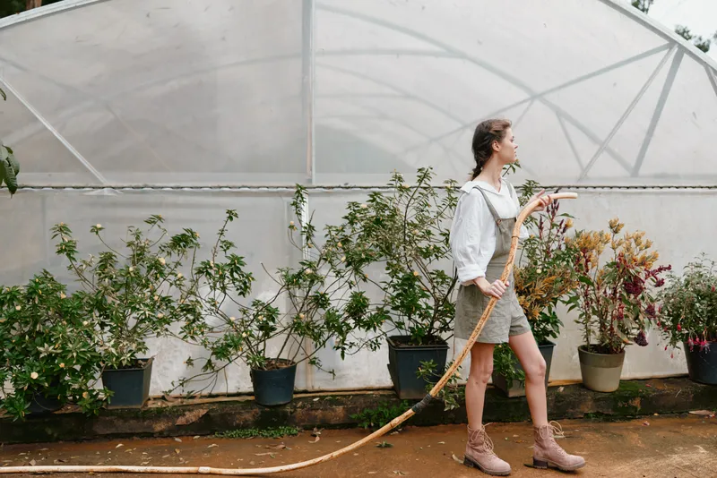 Young woman in stylish overalls watering plants in a greenhouse, showcasing a connection with nature and sustainable living through vibrant greenery and earthy tones