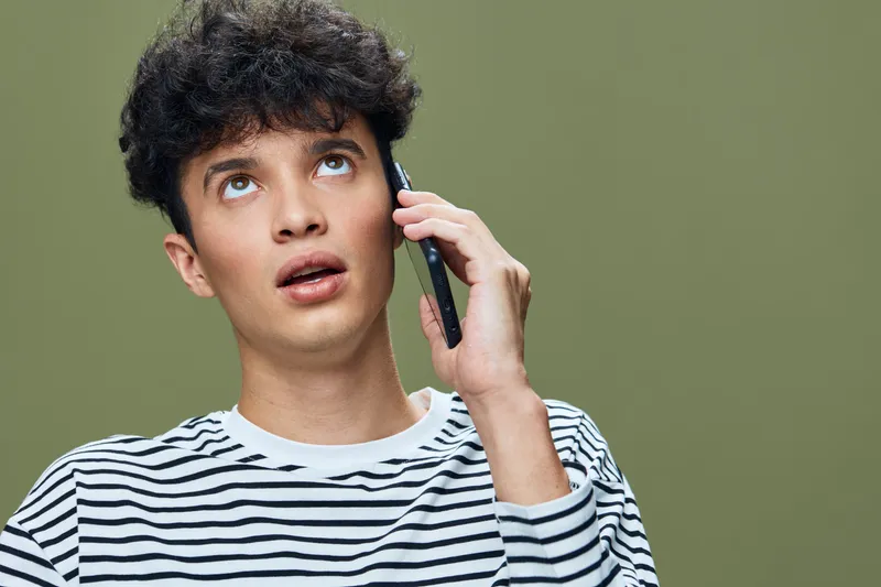 Young man talking on smartphone looking surprised, wearing striped shirt with curly hair against green studio background, casual modern style communication.