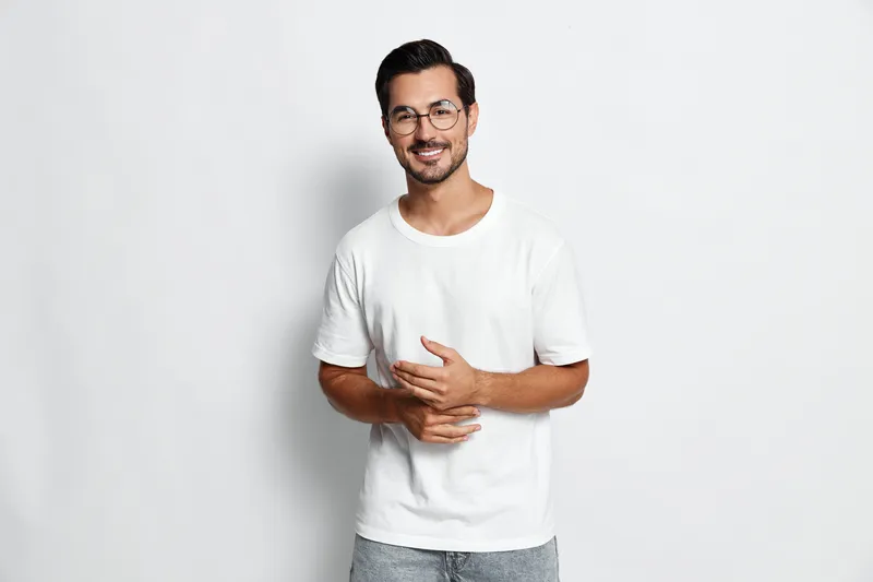 Smiling cheerful young man wearing glasses and casual white t-shirt posing confidently with hands together against plain solid background. People lifestyle concept.
