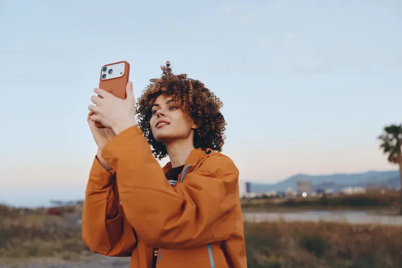Woman lifestyle selfie outdoors, holding phone near face, curly hair, orange jacket, smiling in open field at sunset, soft breeze, natural light, calm mood, distant haze