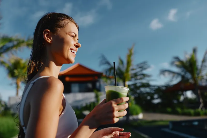 Smiling woman holding a green smoothie in a sunny outdoor setting, relaxed lifestyle, wellness and self care. Bright natural light creates a warm mood and refreshment.