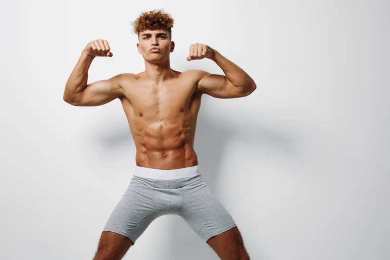 Young muscular man flexing biceps and showing fit body with defined abs wearing gray shorts on white background for fitness and strength concept.