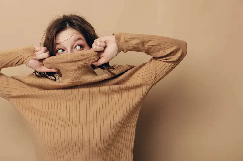 Young woman in brown knitted sweater pulls collar over face, looking sideways with curious expression. Neutral beige background creates warm and cozy atmosphere. Concept of shy or playful mood.