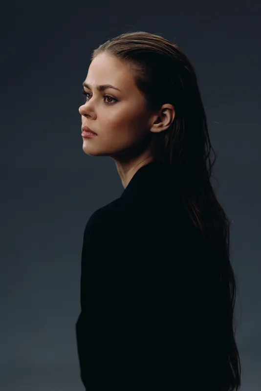 Serious woman in black clothing with side profile against dark background, thoughtful expression, minimalistic portrait style.