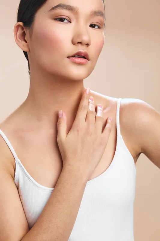 Asian woman in a soft studio portrait wearing a white tank top, delicate makeup, and a serene expression, highlighting natural beauty and calm confidence.