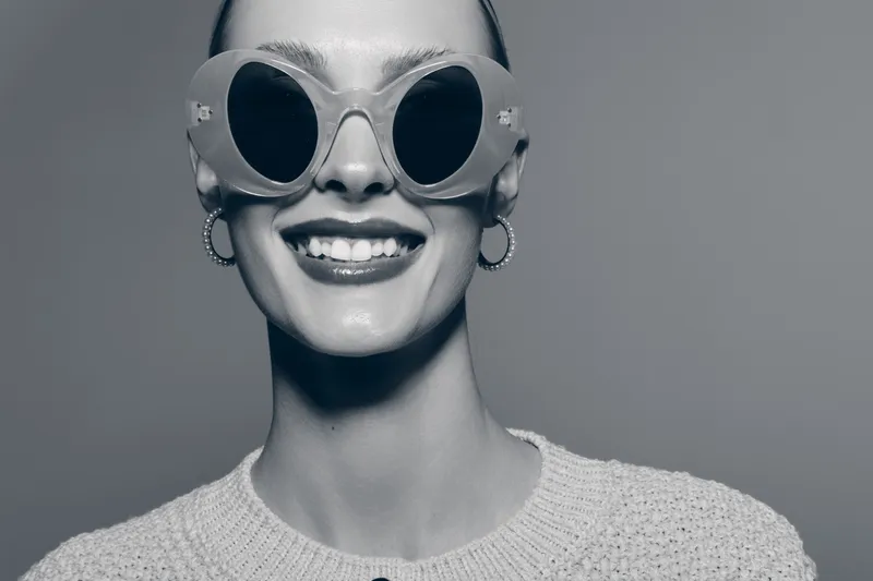 Closeup of smiling young caucasian woman with fair skin wearing large round sunglasses and hoop earrings on grey background. Concept of fashion, style, and positive mood.