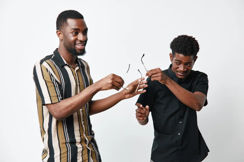 Two young black men with dark skin smiling and interacting while holding eyeglasses in casual clothing on white background. Friendship, happiness and positive mood indoors.