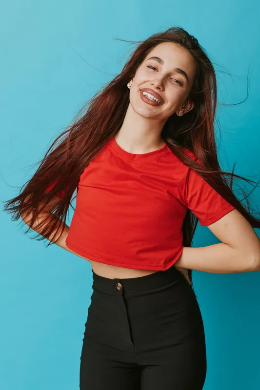Young woman smiling with long hair wearing red crop top and black pants standing against blue background. Happy female model posing with hands on hips and joyful expression.