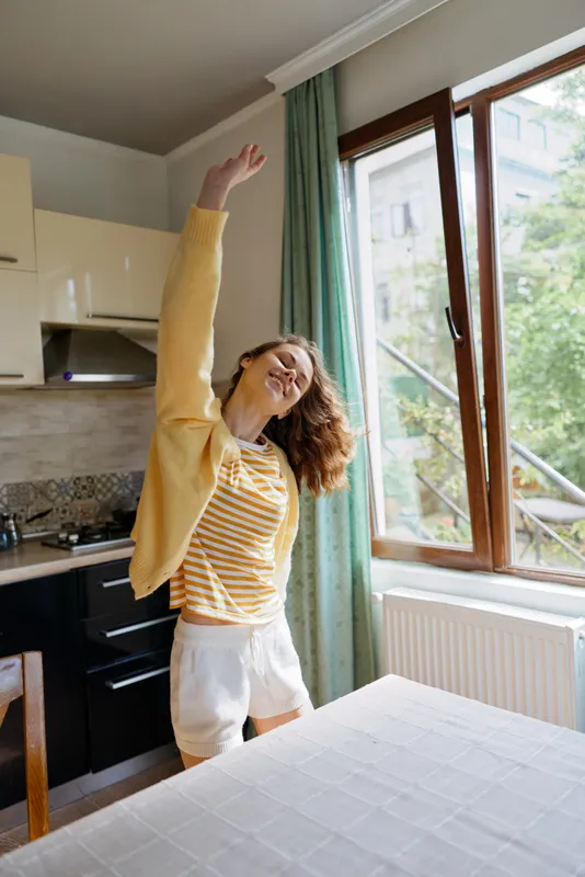 Cheerful woman indoors in a cozy kitchen, stretching with raised arm, striped shirt, soft cardigan, daytime sunlight through a large window, relaxed lifestyle moment, bright colors, home atmosphere,