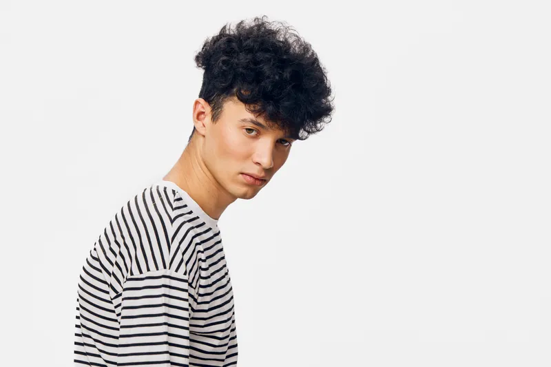 young man with curly hair wearing striped shirt against white background, looking serious with casual style and modern haircut, studio portrait