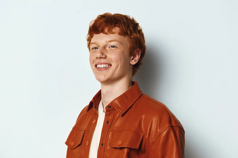 Young man with red hair smiling confidently against a white background. Casual style wearing a brown leather jacket and light shirt for portrait photography session.