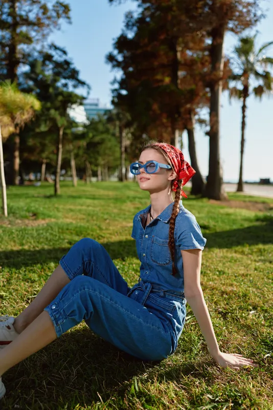 lifestyle woman in modern boho-western denim street style sits on grass outdoors with sunglasses and red bandana, captured in warm film color tones, relaxed mood