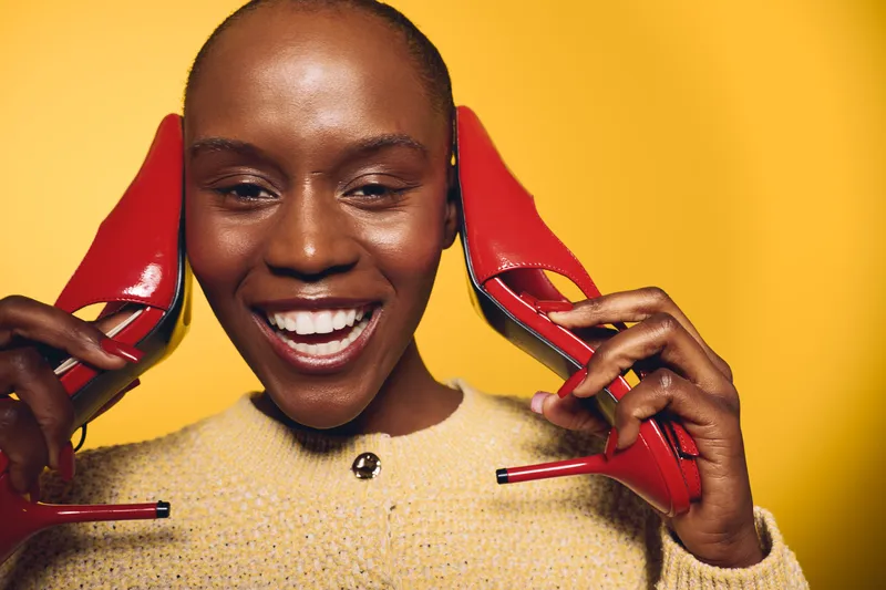 Happy black woman with dark skin holds red high heels near ears smiling against yellow background. Concept for fashion, joy, beauty, and lifestyle with vibrant colors and positive mood.