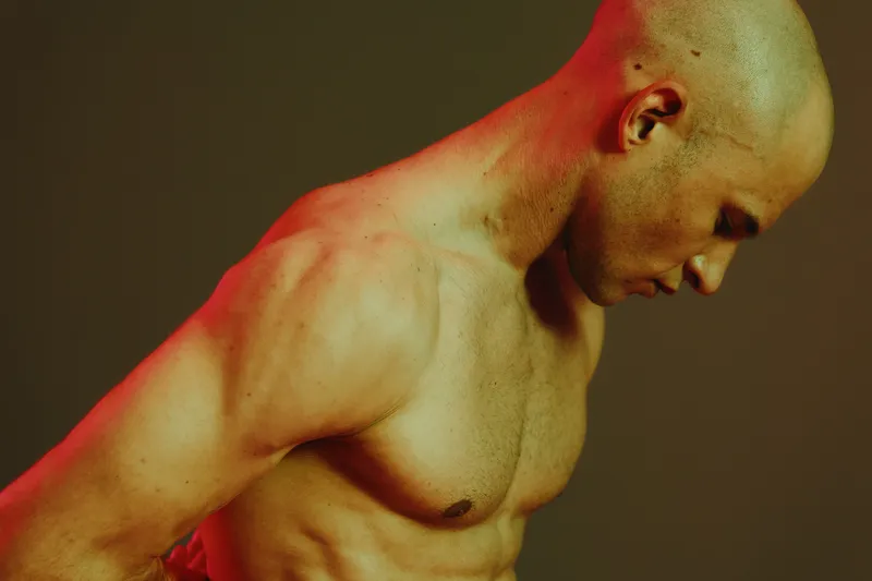 Muscular man in contemplative pose, showcasing emotional depth with dramatic lighting highlighting his physique, against a moody background.