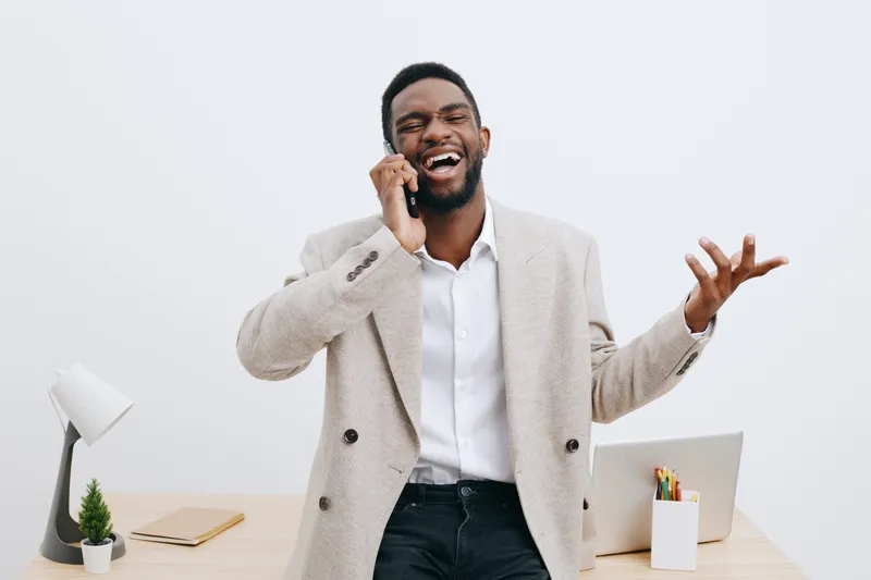 Smiling man in beige blazer talks on mobile phone, looks cheerful and confident, standing in modern office with white background, casual business attire, lifestyle concept.