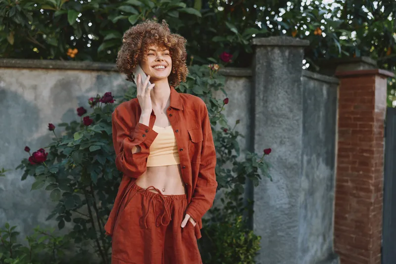 A cheerful woman in a rust jacket stands outdoors, talking on a cellphone, smiling. Bright garden background, casual lifestyle moment captured for fashion and everyday life
