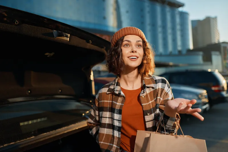 Woman with shopping bag by car trunk smiling in urban golden hour, candid lifestyle moment capturing authenticity, mindful living and emotional storytelling in a modern city scene.
