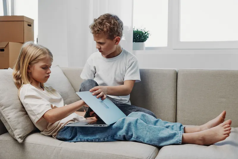 Two children sitting on a beige couch using a laptop together in a bright living room. Casual clothing and natural light create a relaxed atmosphere for learning and collaboration.