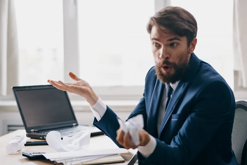Бородатый мужчина работает за ноутбуком в офисе, на его лице недовольство