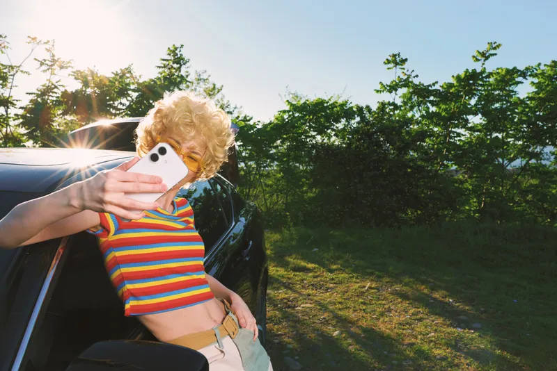 Bright portrait of a young person with curly blonde hair in a rainbow striped top, leaning from a car window to selfie, sunny roadside scene with natural light.