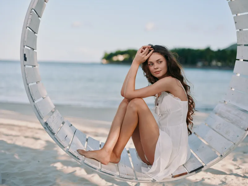 A young woman sits in a white dress on a circular beach swing by the calm sea, warm sand and soft light creating a tranquil coastal portrait.