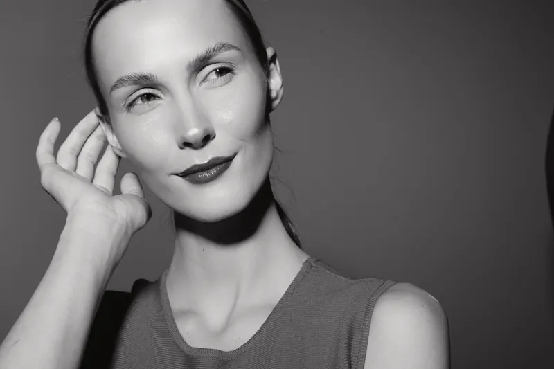 Portrait of a young white woman with fair skin and slicked back hair posing with hand near ear. Soft studio lighting highlights her smooth complexion and subtle expression.